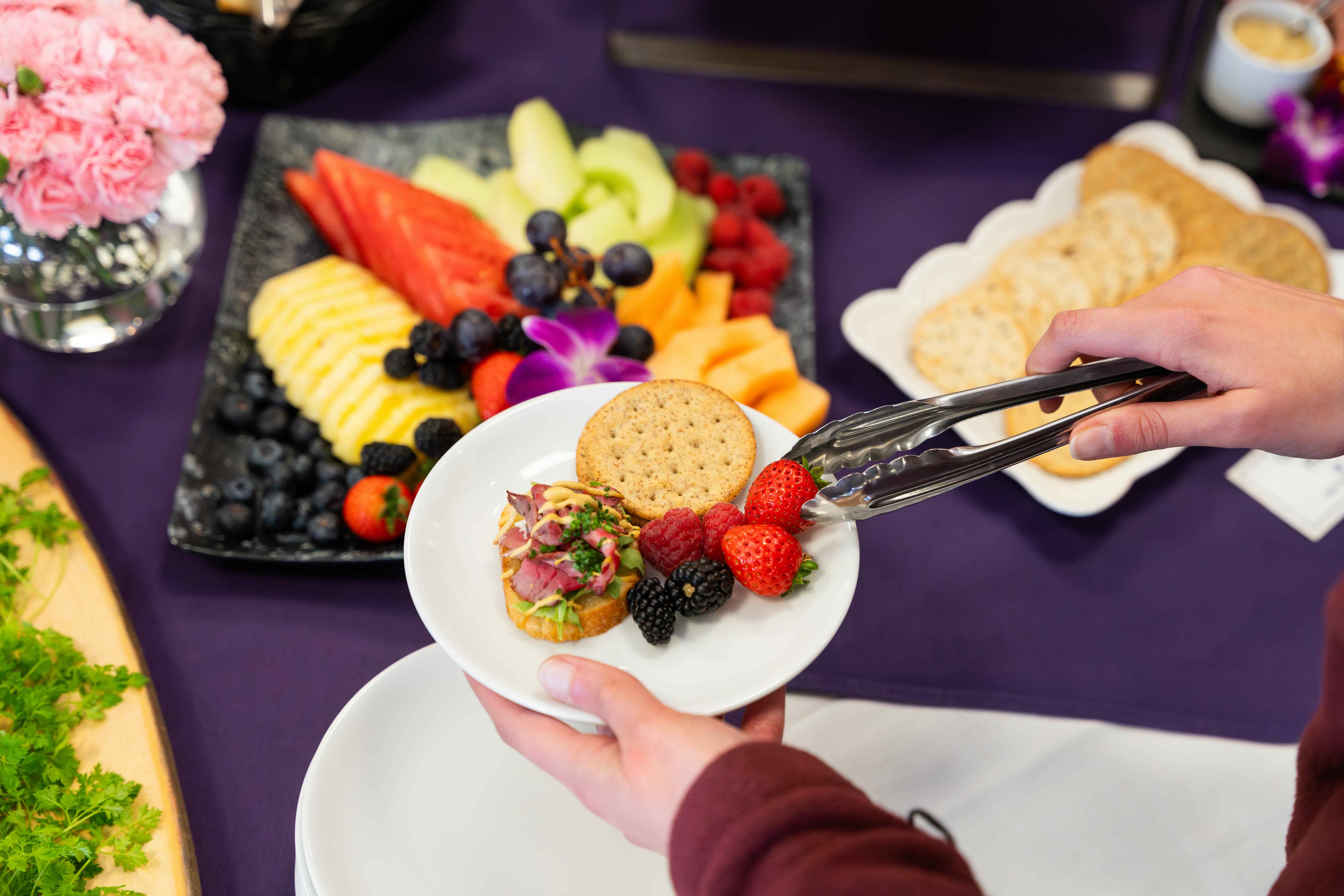 Close-up of a plate with crackers, berries, and an appetizer being served with tongs; fruit platter in the background.