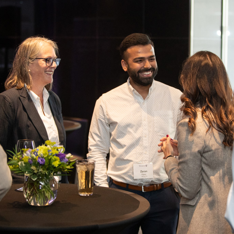 Three people standing around a tall round table with drinks and a small floral centerpiece, engaged in conversation at an indoor networking event.
